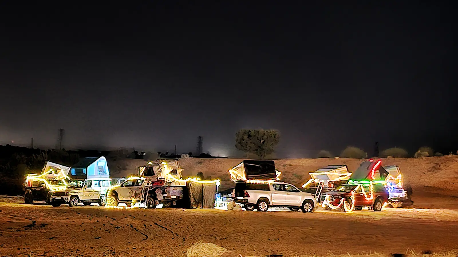 Overlander pickups and SUVs with rooftop tents strung with festive lights at a desert night camp